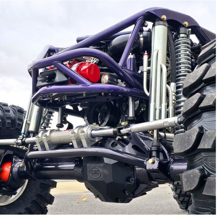 Close-up of a 14 inch travel 3.0 bypass shock mounted on a purple and black off-road vehicle chassis with large off-road tires, silver suspension springs, and a red engine component.