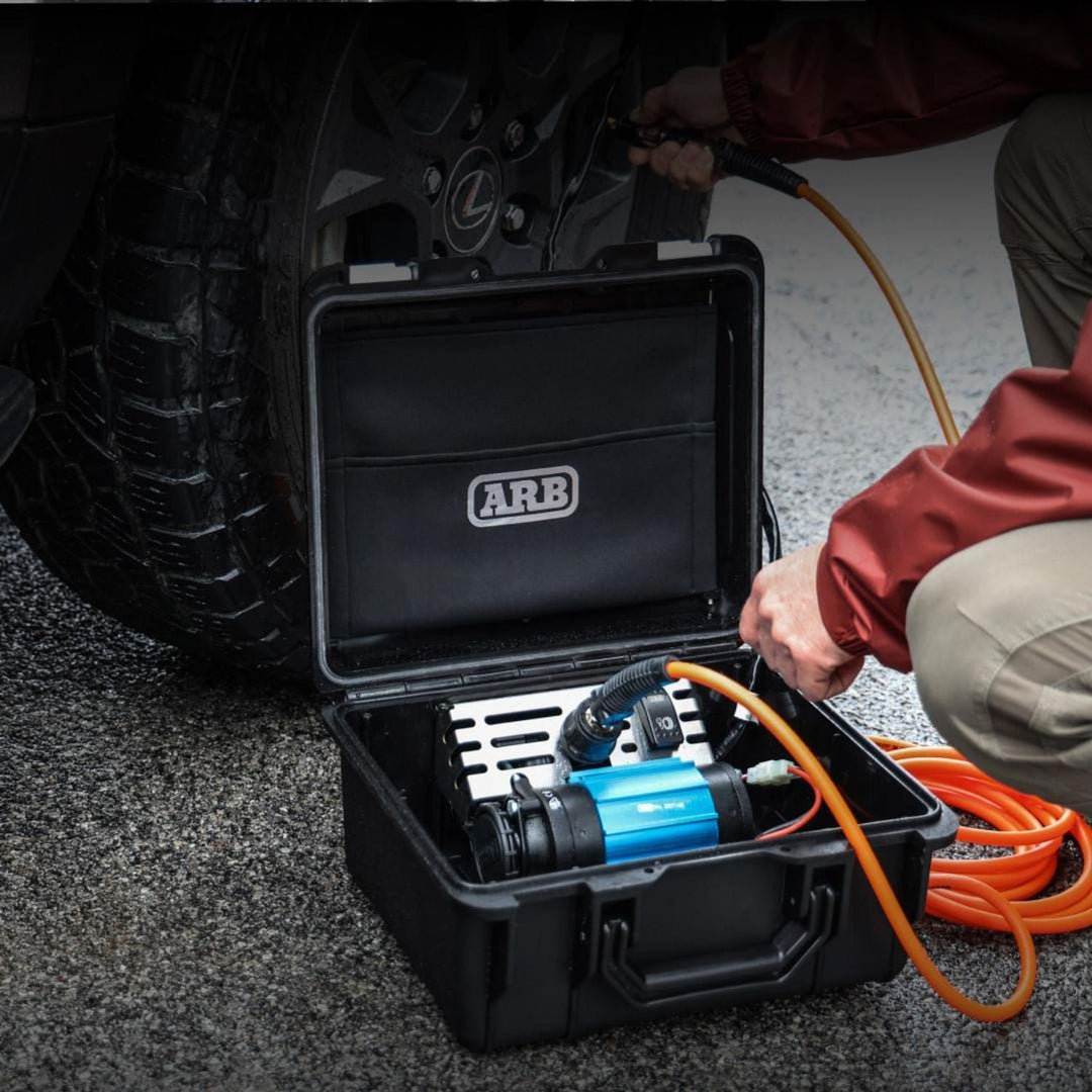 Black weatherproof portable compressor for overlanding with ARB logo open, in use, on a car tire.